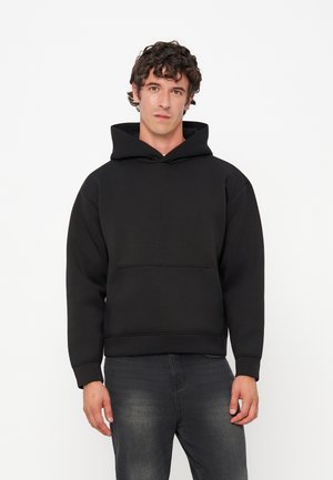 Homme aux cheveux bouclés portant un sweat à capuche noir et un jean foncé, debout devant un fond blanc uni.