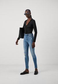 Black long-sleeve bodysuit with gathered detail, blue skinny jeans with distressed knees, black tote bag, and black slip-on shoes.