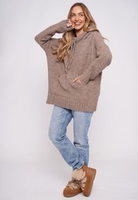 Khaki, oversize'owy, dziergany sweter z prążkowanym wykończeniem i ściągaczem, w połączeniu z niebieskimi dżinsami oraz brązowymi sneakersami z wyściółką ze skóry owczej.