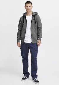 Grå zip-up hættetrøje med lommer og sorte snore, parret med marineblå cargo bukser med sidelommer, og sorte sneakers med hvide såler.