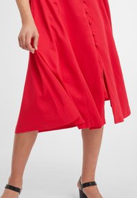 Robe rouge, de longueur genou, faite d'un tissu lisse avec des boutons sur le devant et une fente sur le côté. Portée avec des sandales à talons hauts noires.