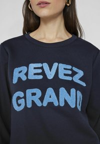 Tamsiai mėlynas džemperis su tekstūriniu šviesiai mėlynu užrašu "REVEZ GRAND" priekyje. Apvalus kaklas ir laisvas siluetas.