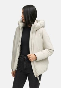 Donna con capelli neri lisci che indossa un piumino beige chiaro sopra una maglia nera e jeans neri, in piedi contro uno sfondo semplice.