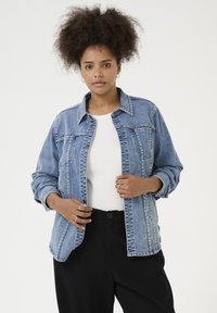Chaqueta de mezclilla con acentos de tachuelas, color azul y un cuello clásico. Presenta dos bolsillos en el pecho y cierre de botones, combinada con una blusa blanca.