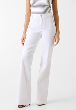 Γυναίκα που φοράει λευκό παντελόνι καμπάνα με ψηλή μέση και χρυσά κουμπιά στα πλάγια, συνδυασμένο με αμάνικη ριγέ μπλούζα και καφέ σανδάλια.