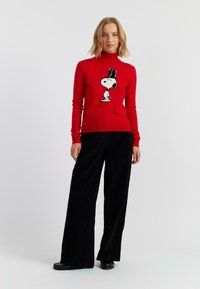 Rode coltrui met een Snoopy-afbeelding, gecombineerd met zwarte wijde broek. De trui heeft ribgebreide manchetten en zoom, met een gladde gebreide textuur.