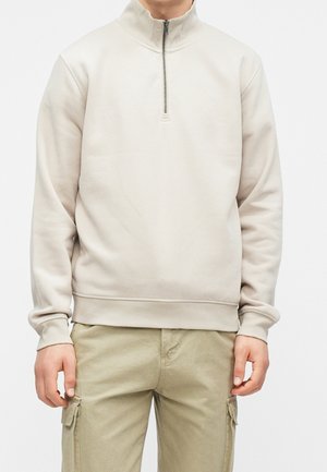 Personne portant un pull beige clair à fermeture éclair sur le quart et un pantalon cargo kaki, debout, les bras détendus le long du corps.