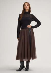Top nero a maniche lunghe con texture in rete, abbinato a una gonna in tulle marrone fluida con bordo in vita nera e dettaglio in nastro. Stivali neri alla caviglia.