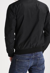 Czarna lekka kurtka bombowiec o gładkiej fakturze i ściągaczowych mankietach, w zestawieniu z ciemnoniebieskimi dżinsami z delikatnymi detalami przeszyć.