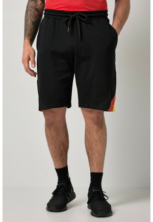 Homme portant un short de sport noir avec des accents rouges et jaunes, des chaussettes noires, et des baskets noires, debout, les mains détendues.