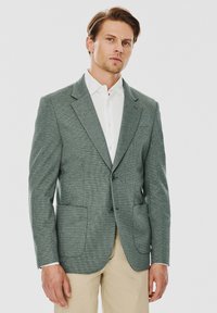 Jasnozielona żakiet o teksturze z klapami w kształcie litery notch, z dwoma guzikami i dwiema przednimi kieszeniami. Połączony z białą koszulą z kołnierzykiem oraz beżowymi spodniami.