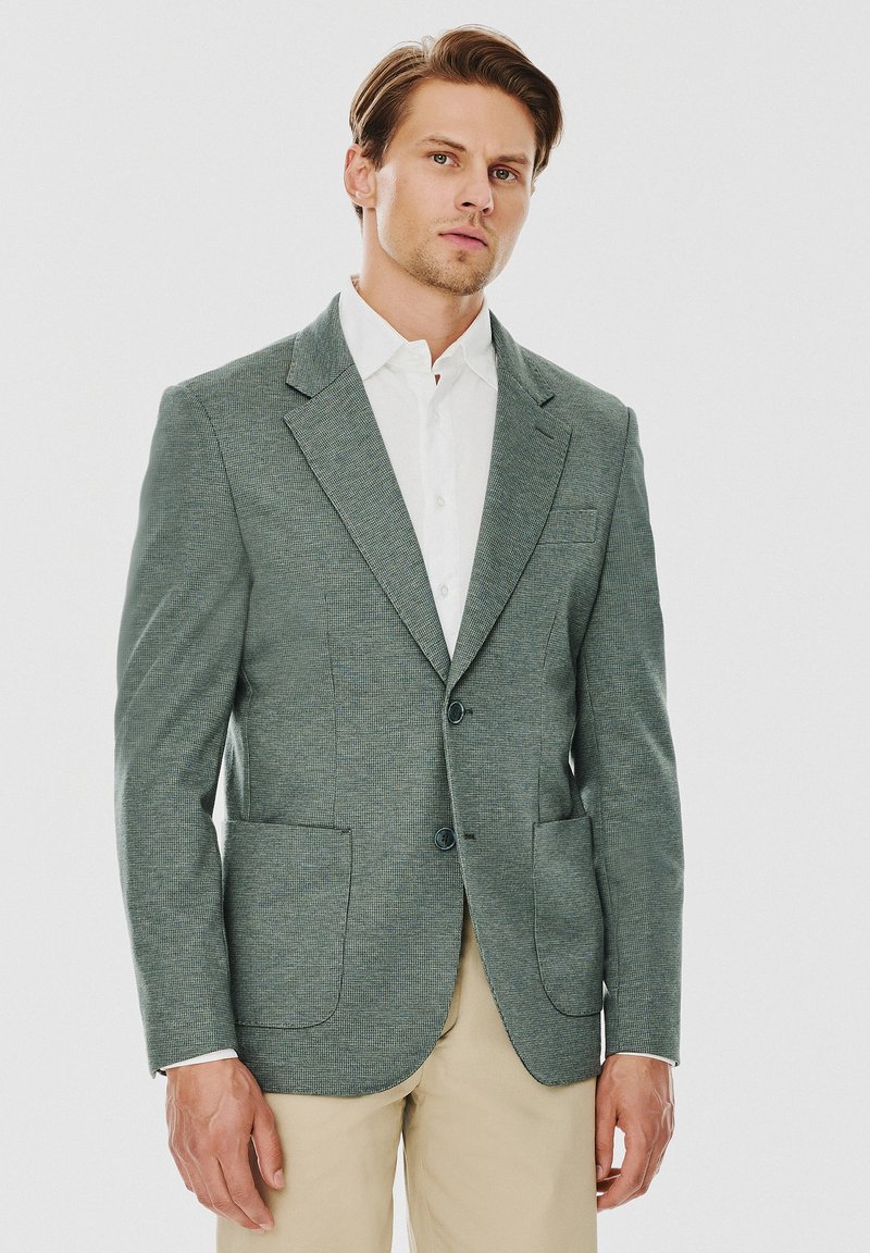 Jasnozielona żakiet o teksturze z klapami w kształcie litery notch, z dwoma guzikami i dwiema przednimi kieszeniami. Połączony z białą koszulą z kołnierzykiem oraz beżowymi spodniami.