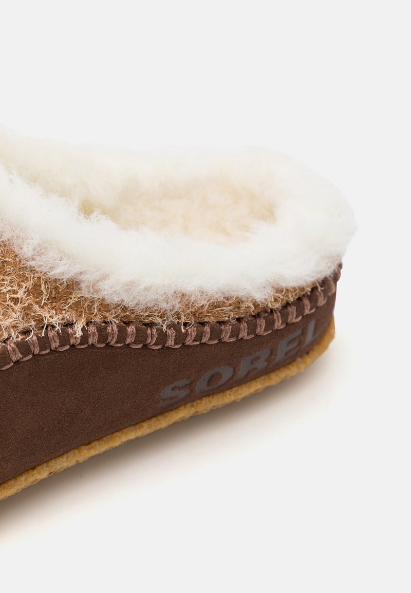 Bruine slipper met een zachte, crèmekleurige pluche voering. Heeft een getextureerde buitenkant en gestikte accenten. Merknnaam "SOREL" zichtbaar.