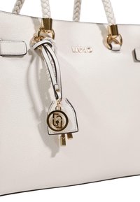 Sac à main en cuir blanc texturé avec poignées tressées, ferrures dorées et une étiquette décorative avec un logo et de petits charms.