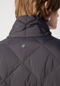 Gequilted donkergrijze jas met een hoge kraag en een metalen logo-accent nabij de halslijn. Gladde, gestructureerde stof met een ruitpatroon.