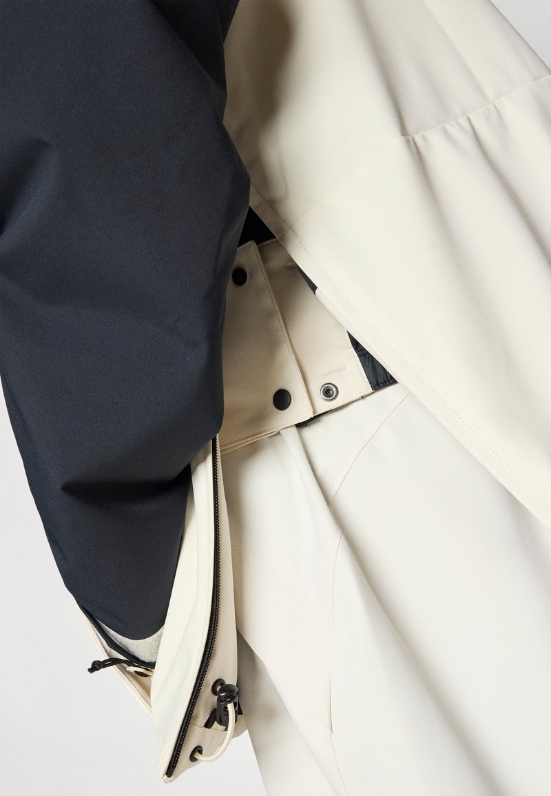Veste imperméable avec une partie supérieure marine et une partie inférieure crème, dotée d'une fermeture éclair, de boutons-pression et d'un cordon de serrage à l'ourlet.