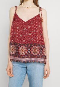 Top a canotta rosso floreale con spalline regolabili e un orlo decorato in navy, crema e accenti dorati. Tessuto leggero con una vestibilità ampia.