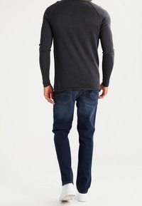 Maglietta grigia a maniche lunghe con maniche raglan abbinata a jeans blu scuro. Modello che indossa sneakers bianche, visto da dietro.