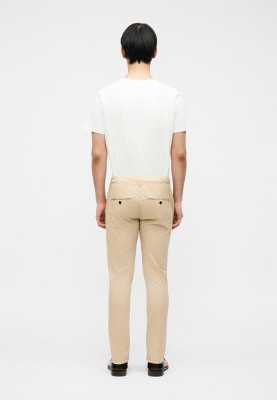 Άτομο που στέκεται γυρισμένο προς τα πίσω, φοράει άσπρο μπλουζάκι, μπεζ παντελόνι με πίσω τσέπες και μαύρα παπούτσια, μπροστά σε απλό ανοιχτό φόντο.