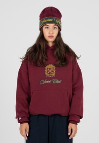 Burgundowa bluza z kapturem z przednią kieszenią, ozdobiona haftowanym herbem i napisem "Social Club" w niebieskim i żółtym kolorze. W komplecie z pasującą czapką.
