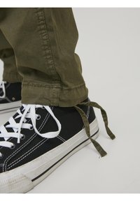 Spodnie z zielonego oliwkowego twillu z podwiniętymi mankietami, zestawione z czarnymi tenisówkami z płótna z białymi sznurówkami i białą gumową podeszwą.