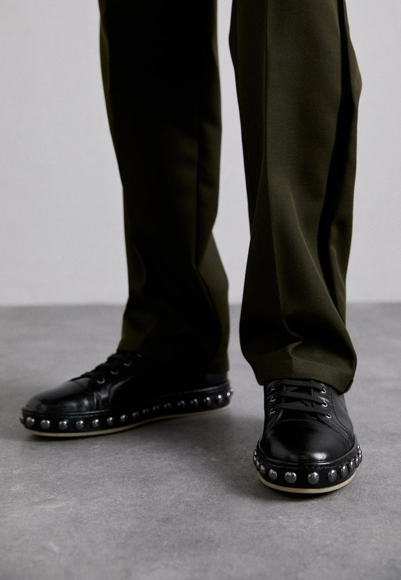 Black leather sneakers with a rounded toe, silver studs along the sole, and black laces. Paired with dark olive green pants.
