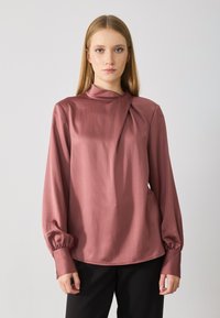 Donna con capelli biondi lisci indossa una blusa a maniche lunghe color rosa polveroso con scollatura plissettata e pantaloni neri, in piedi su uno sfondo chiaro e semplice.