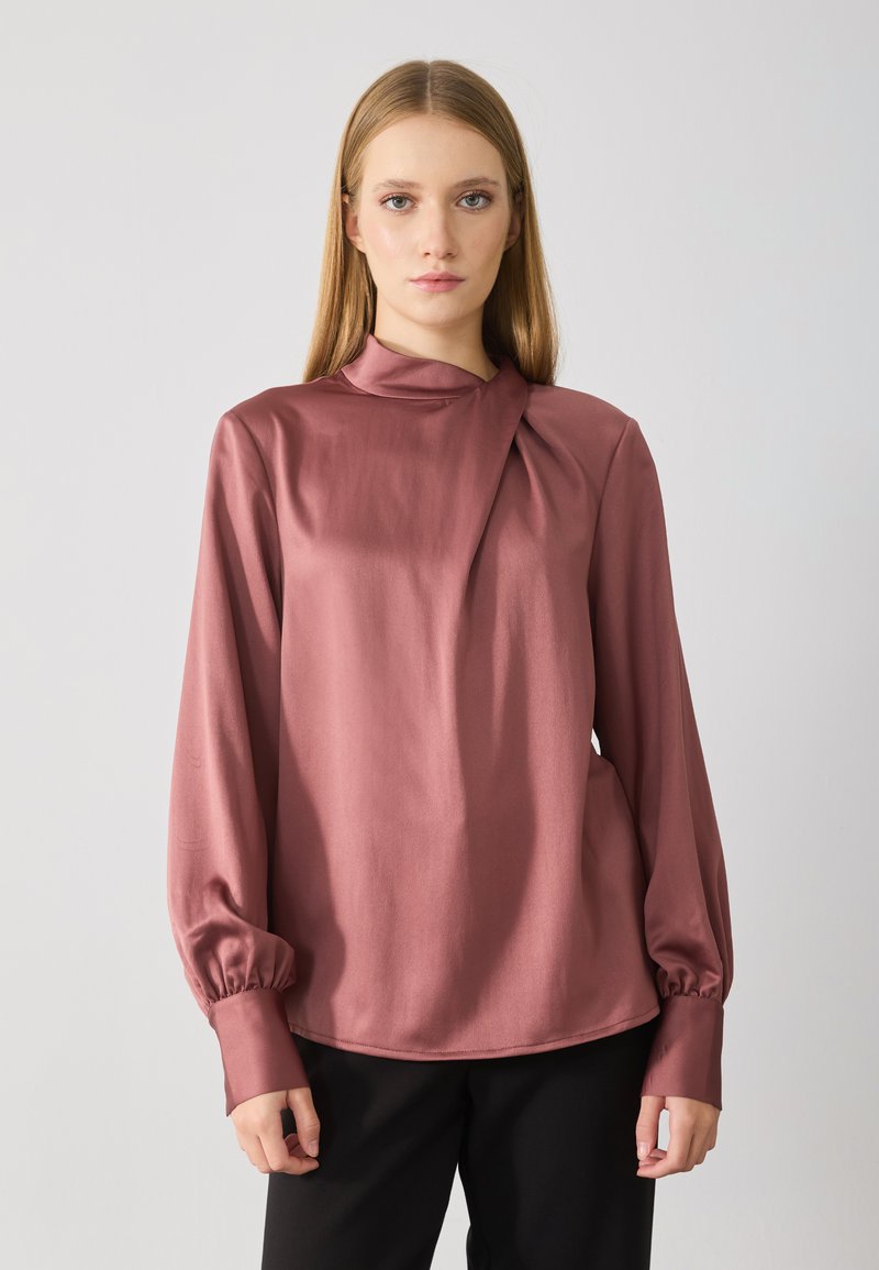 Donna con capelli biondi lisci indossa una blusa a maniche lunghe color rosa polveroso con scollatura plissettata e pantaloni neri, in piedi su uno sfondo chiaro e semplice.