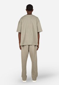 Osoba stojąca plecami, ubrana w luźny beżowy top z krótkim rękawem w prążki oraz pasujące szerokie spodnie, z białymi butami na białym tle.