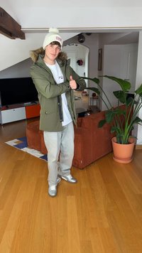 Zelena parka z rjavim krznom obrobljeno kapuco, bela kapa z logotipom, bela majica, svetle kavbojke in srebrni športni copati v dnevni sobi.