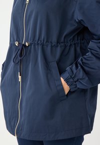 Giacca con zip di colore blu navy, con polsini elasticizzati, vita regolabile con cordino e tasche laterali nascoste, realizzata in un tessuto leggero e morbido.