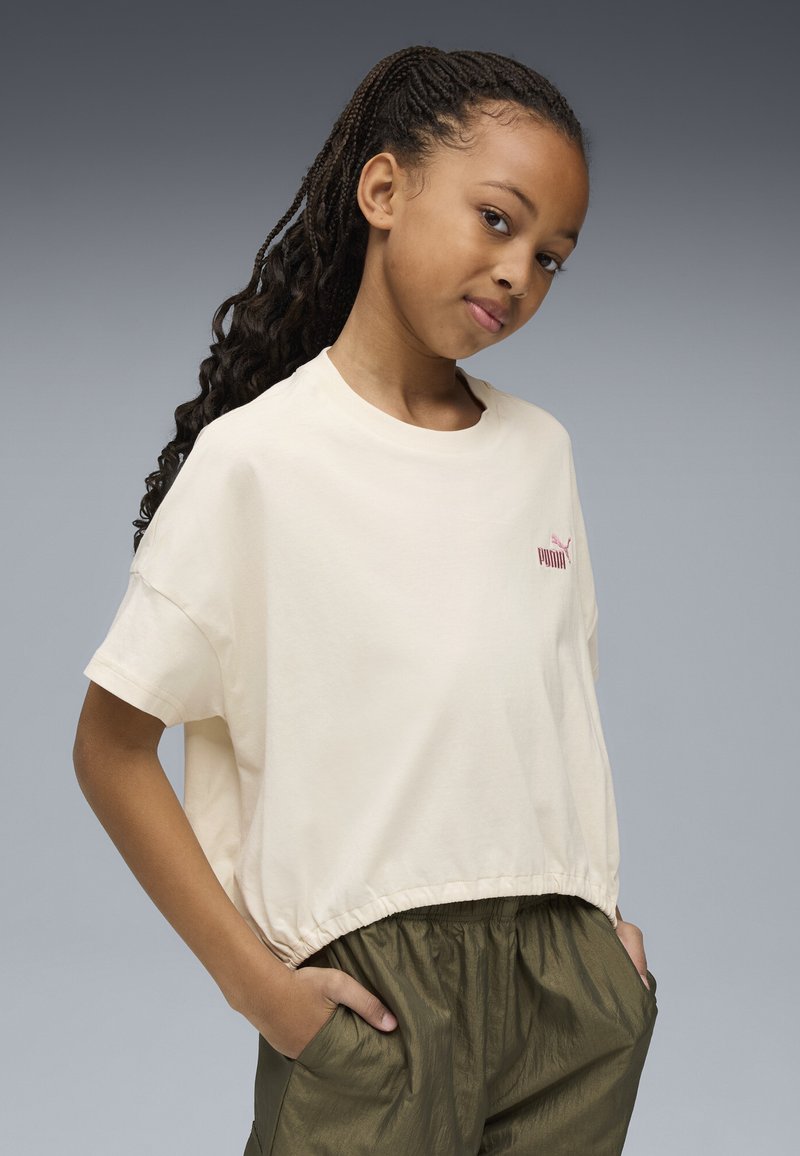 Niña joven con el cabello largo trenzado, vistiendo una camiseta crema de Puma y pantalones verde oliva, de pie con las manos en los bolsillos frente a un fondo gris.