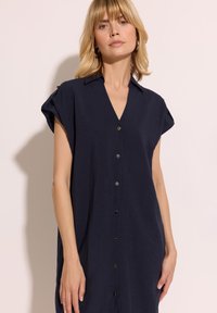 Granatowa sukienka zapinana na guziki z dekoltem w kształcie litery V, krótkimi rękawami i metalowymi detalami guzików. Wykonana z miękkiego, lekkiego materiału o luźnym kroju.