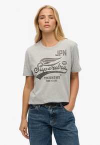 T-shirt en coton gris avec un design graphique comprenant le texte "Superdry" et un accent d'aile, à manches courtes et à coupe décontractée. Porté avec un jean.