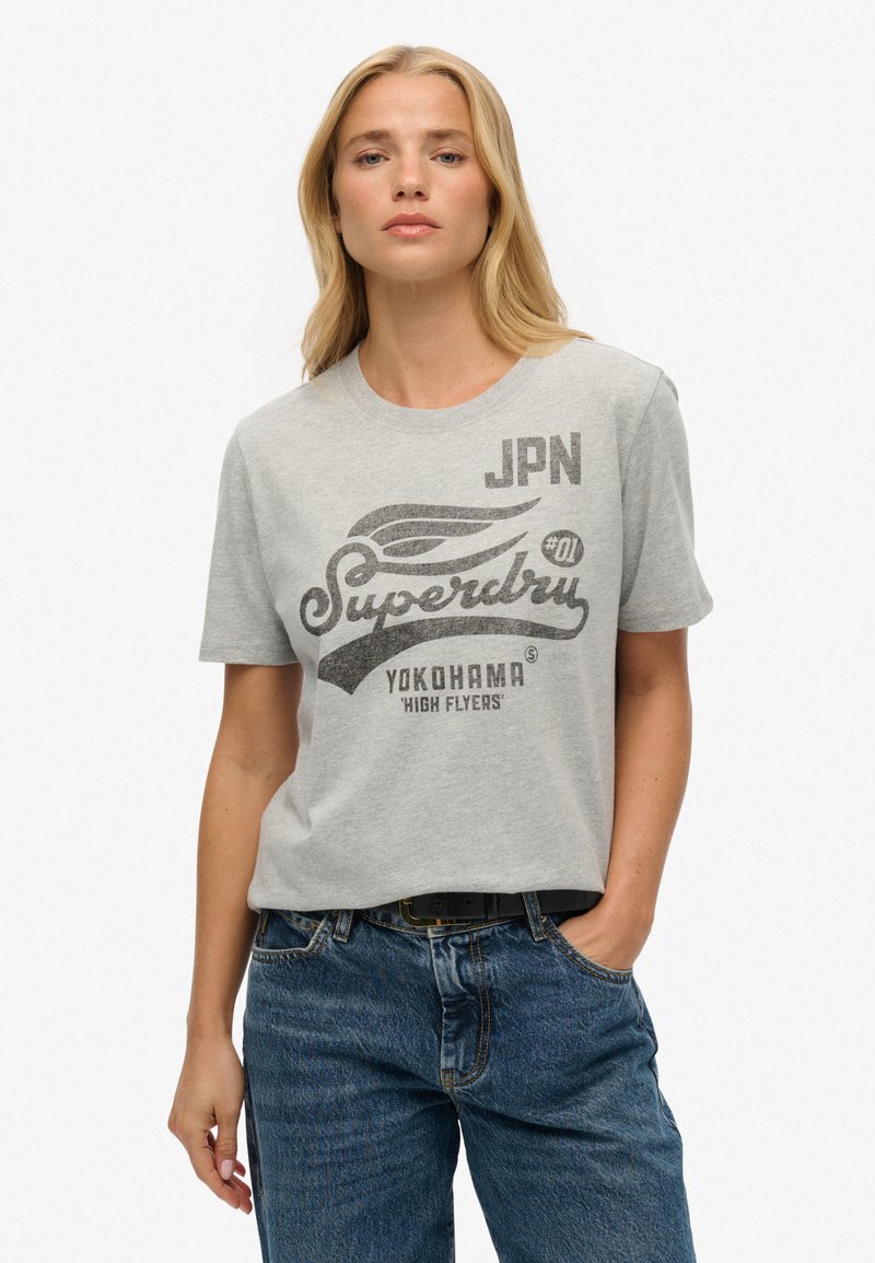 T-shirt en coton gris avec un design graphique comprenant le texte "Superdry" et un accent d'aile, à manches courtes et à coupe décontractée. Porté avec un jean.