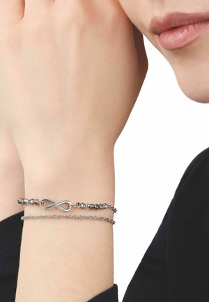 Muñeca con dos pulseras plateadas, una con un símbolo de infinito y pequeñas cuentas, frente al rostro de una persona y una manga negra.