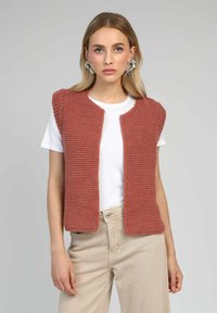 Gilet sans manches en tricot couleur rose poussiére, présentant un motif texturé, une ouverture à l'avant et un col arrondi, porté avec un T-shirt blanc.