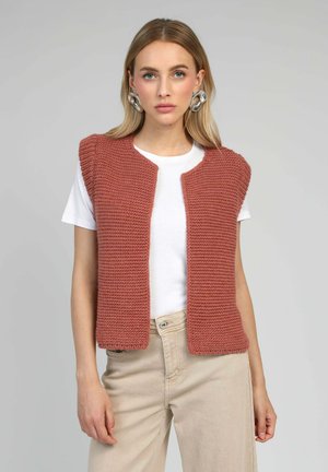 Gilet sans manches en tricot couleur rose poussiére, présentant un motif texturé, une ouverture à l'avant et un col arrondi, porté avec un T-shirt blanc.