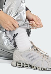 Γκρι αθλητικά παπούτσια με υλικά από δίχτυ και σουέντ, με χοντρή σόλα. Λευκές κάλτσες με μαύρο λογότυπο. Ορατά μεταλλικά ασημί παντελόνια φόρμας.