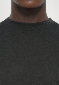 Nærbilde av en person som har på seg en mørkegrå genser med rund hals, med subtil preget tekst på kragen.