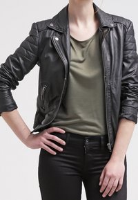 Giacca da moto nera in pelle con dettagli trapuntati sulle spalle, abbinata a una t-shirt verde e jeans neri, che mettono in risalto una silhouette aderente.