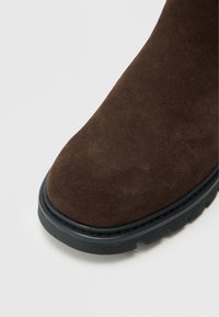 Ruskeat mokkasinilkkurit pyöreällä kärjellä, mustalla kumipohjalla ja teksturoidulla pinnalla. Pohjan reunalla näkyy tikattu sauma.