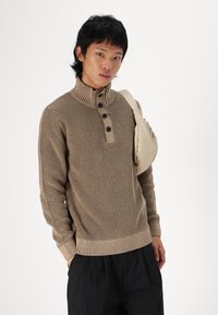 Beżowy sweter z dzianiny z wysokim kołnierzem, trzema czarnymi guzikami, prążkowanymi mankietami i dołem. Modelka nosi jasną torbę na jednym ramieniu.