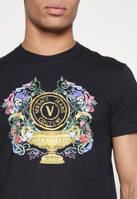 Musta puuvillapaita, jossa on monivärinen kukkadesign ja keskellä kultainen emblemiteksti "VERSACE JEANS COUTURE".
