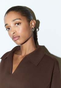 Haut marron à col en V avec une texture lisse. Le modèle porte des boucles d'oreilles en argent circulaires, sur un fond blanc.