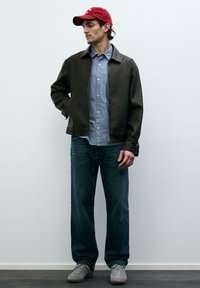 Młody mężczyzna w czerwonej czapce, czarnej kurtce, niebieskiej koszuli w paski, luźnych dżinsach i szarych trampkach, stojący na tle jednolitej białej ściany.