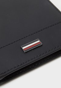Navy leather wallet featuring a rectangular metal logo with red, white, and blue stripes. Smooth texture with visible stitching details.