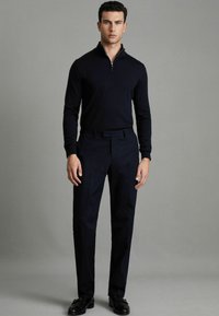 Pull en tricot zippé bleu marine à manches longues, associé à un pantalon sombre taillé et des chaussures noires. La tenue présente une coupe épurée et moderne.