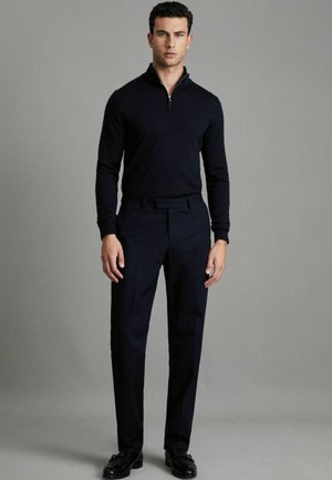 Maglione a zip in maglia di colore navy con maniche lunghe, abbinato a pantaloni scuri sartoriali e scarpe nere. L'outfit presenta una vestibilità moderna e slanciata.