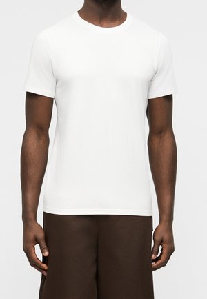 Mann trägt ein schlichtes weißes T-Shirt mit kurzen Ärmeln und dunkelbraune Hose vor hellem Hintergrund.
